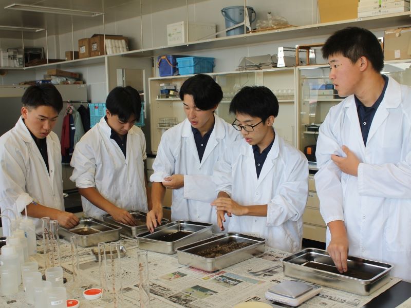 白衣を着た５名の学生が農業改良普及センターで土壌診断実習に取り組む様子