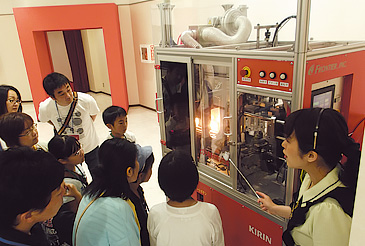 世界初公開となる、透明なボトル型を使用した「ブロー成形機」(左)。素材が一瞬で膨らんでペットボトルへと変わる様子が、はっきりと分かる。