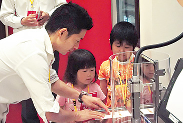 世界初公開となる、透明なボトル型を使用した「ブロー成形機」(左)。素材が一瞬で膨らんでペットボトルへと変わる様子が、はっきりと分かる。