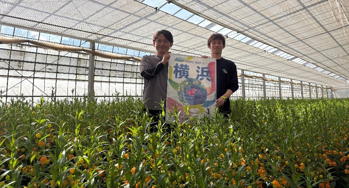 サンダーソニアが育つハウス内で２０２７年国際園芸博覧会のポスターを持つ生産者と丸朝園芸の組合職員
